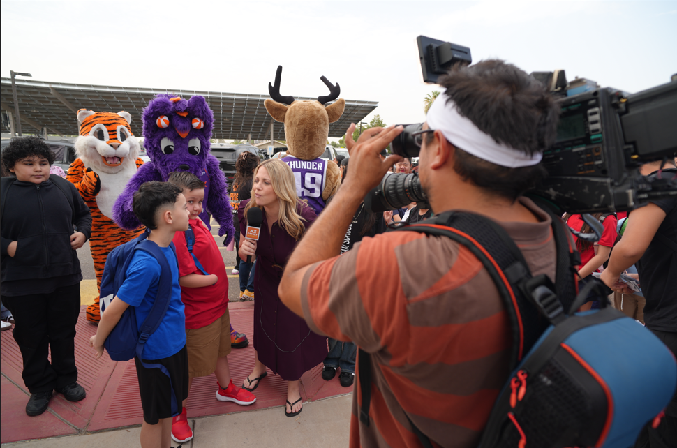 A female reporter interviewing a group of elementary age students outside of school as a cameraman records them