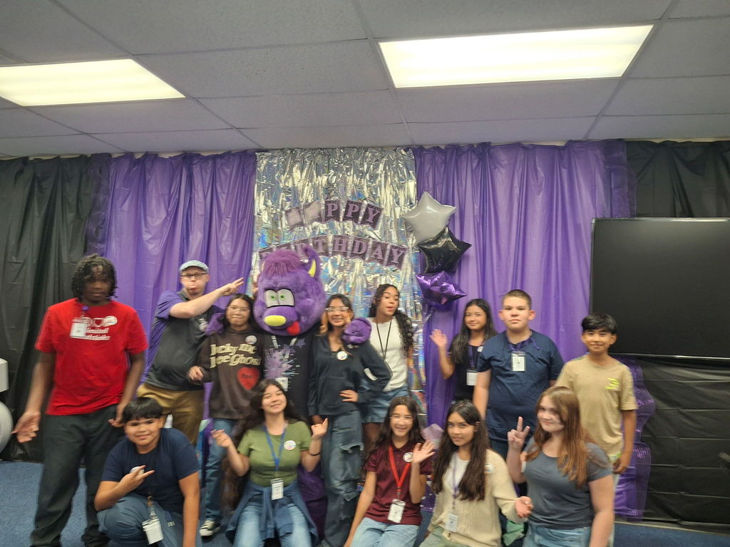 Estrella Middle School students and staff posing together with a purple birthday mascot in front of a decorated backdrop with balloons and a Happy Birthday banner