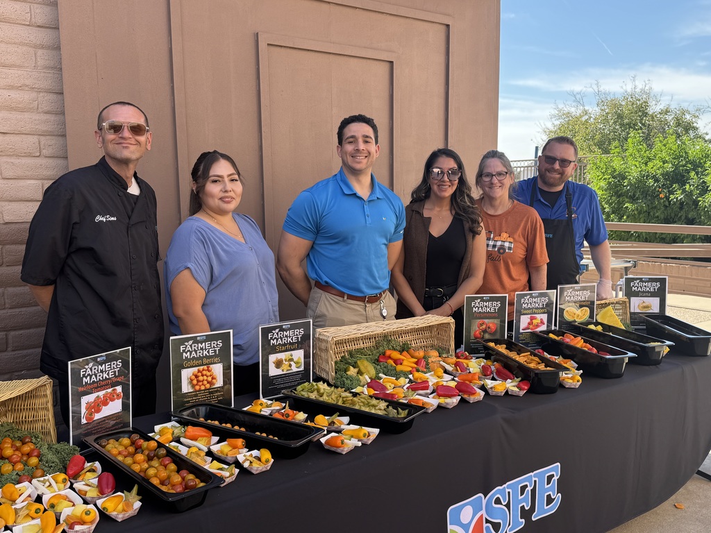 Table full of cherry/grape tomatoes, golden berries, starfruit, persimmons, sweet peppers, and yellow watermelon. Staff smiling behind the table