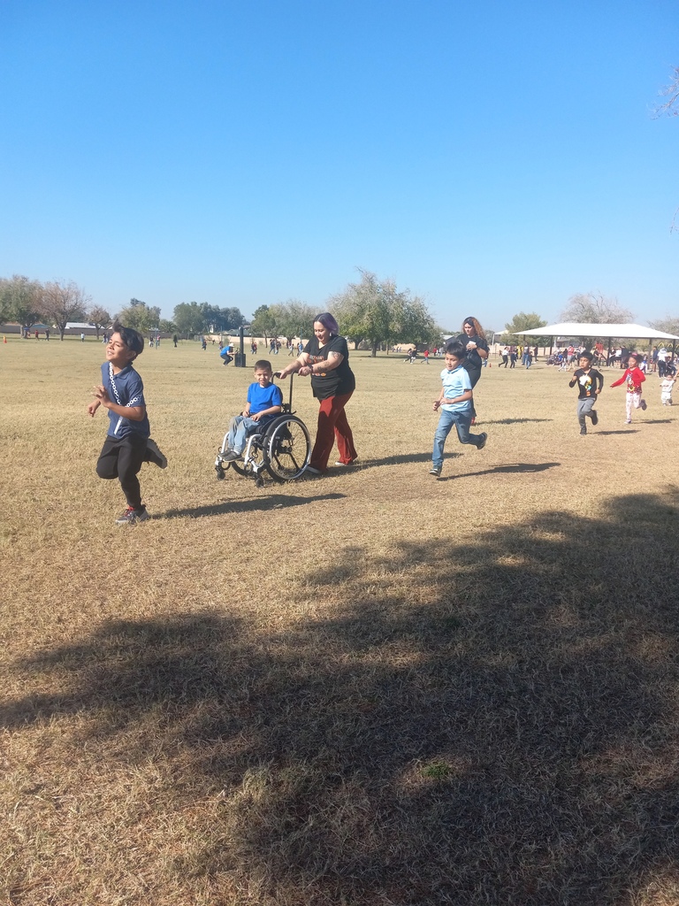 Students running. A student in a wheelchair is being pushed by his teacher around the big grass field.