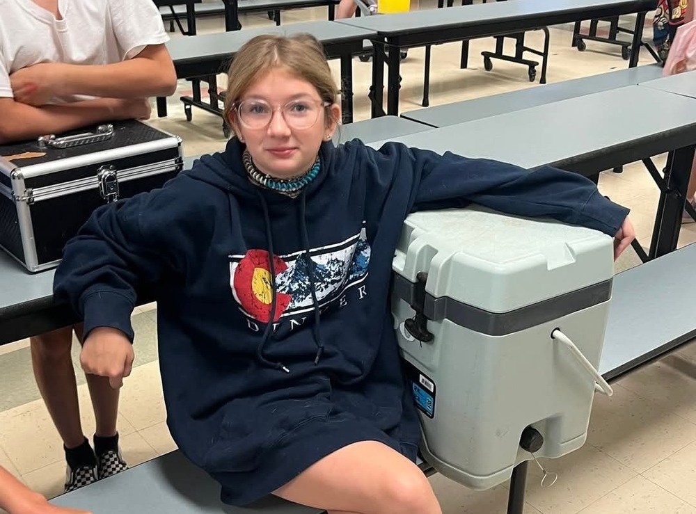 Elementary aged girl with blonde hair and glasses sitting with a gray cooler