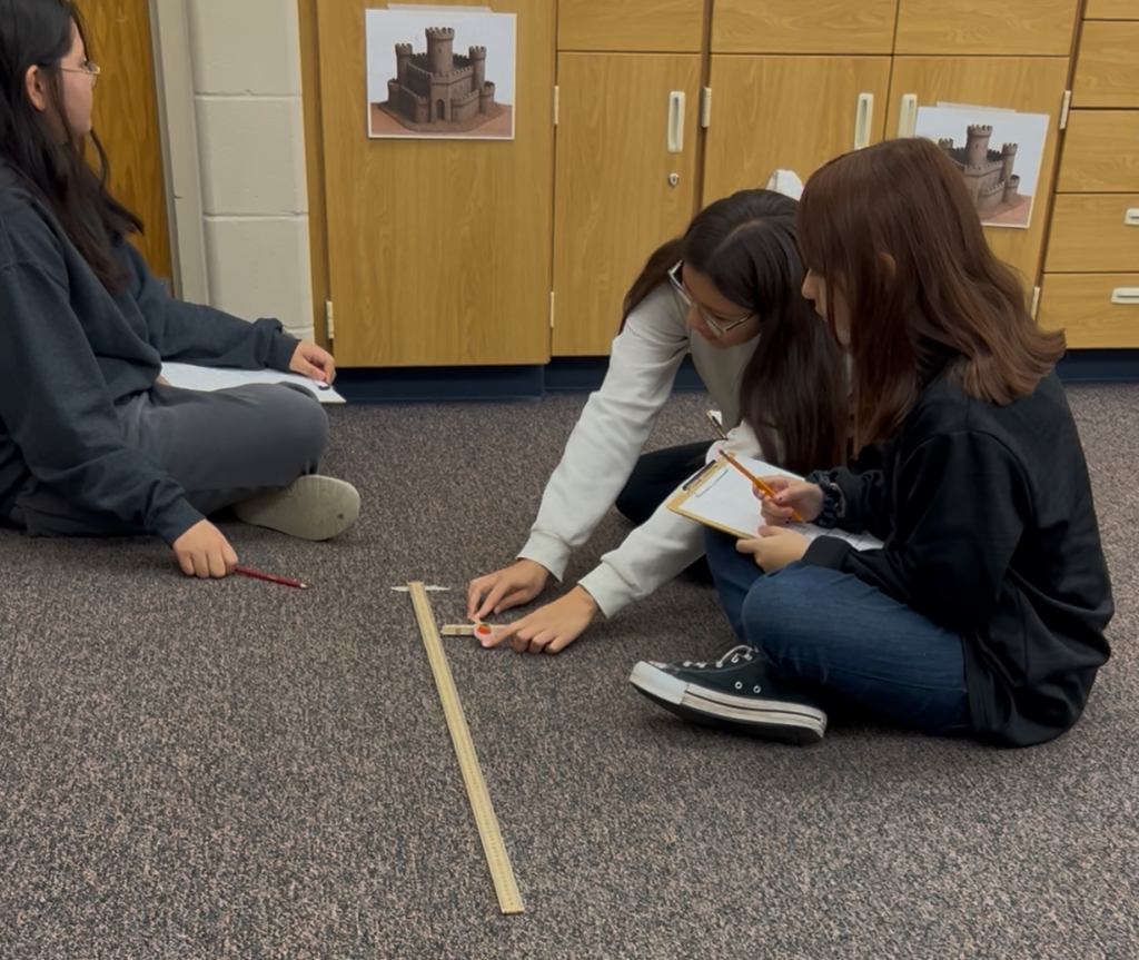Two students watching one student launch a candy pumpkin with a popsicle stick catapult.