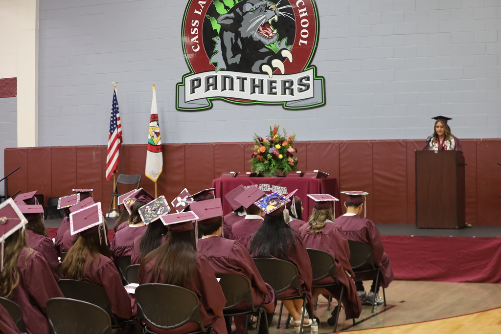 A photo graduates with a stage and podium with flowers, the school logo on the wall and the US and Leech Lake Band of Ojibwe flags.