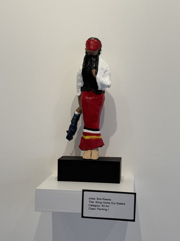 Sculpture of indigenous woman with red ribbon skirt and white shall. Face is covered in red fabric and holding onto a disembodied arm in blue clothing.