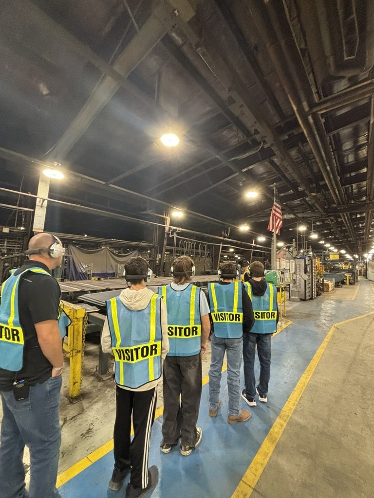 Five students and one teacher all dressed in safety vests looking at production line