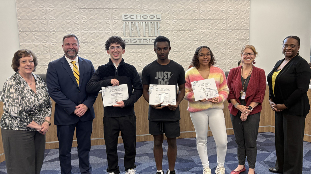Board of Education President, Superintendent, April Yellowjackets of the Month, and Board Vice President stand in front of the board dias.