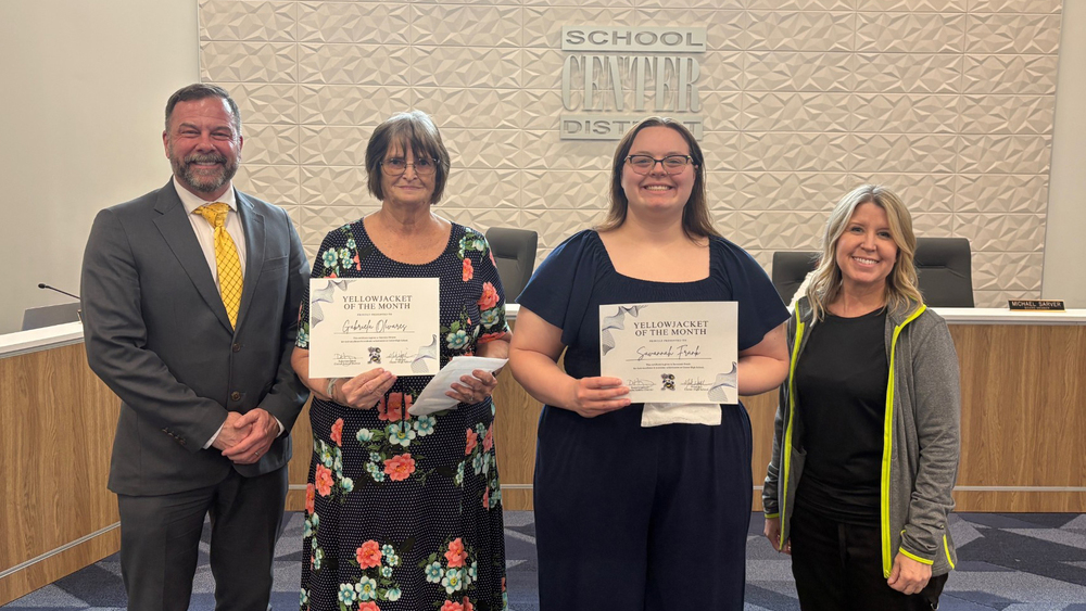 Superintendent Dr. Troy Hogg, Yellowjacket of the Month recipients, and Board of Education Chair Marcie Calvin stand in front of the board dias.