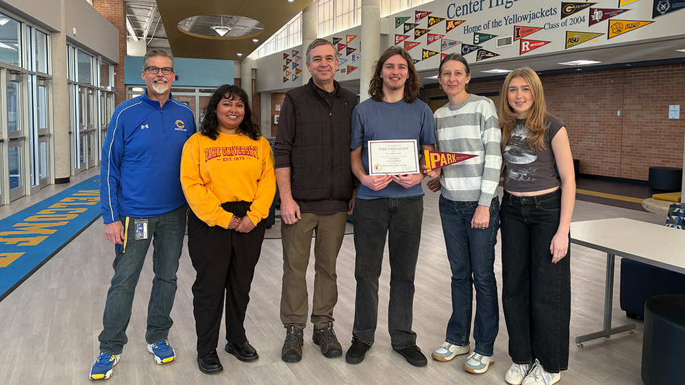 Ezra Van Riette, his parents, sister, and Principal Mark Wiegers, stand in front of the college pennants at Center High School, with a scholarship representative from Park University.