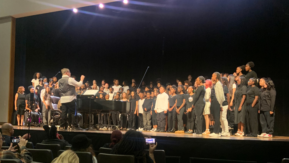 Large group of students stand on stage with bright lights overhead.