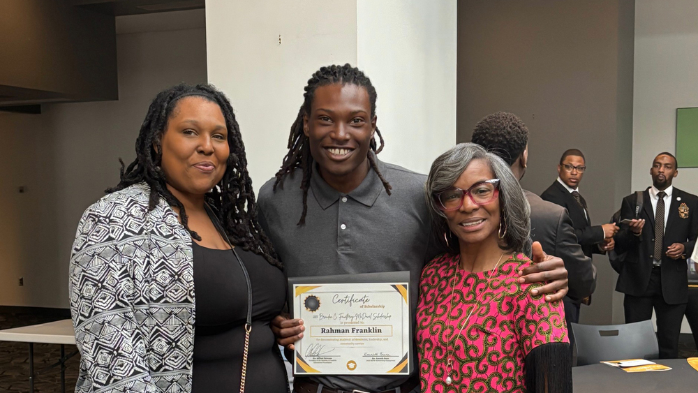 Three people stand indoors; the person in the middle holds a certificate and smiles, while the two people beside them also smile at the camera.