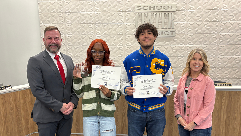 Superintendent Dr. Troy Hogg, student Jade Gray, student Logan Urias, and Board of Education Chair Marcie Calvin stand in a row at the front the boardroom.