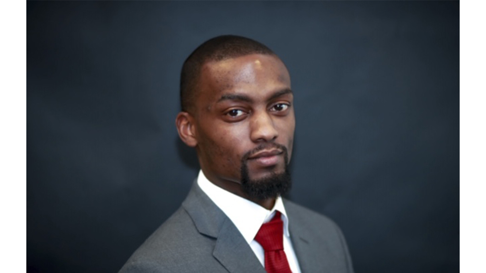 Man in a light-gray suit and dark tie, smiling and looking directly at the camera.