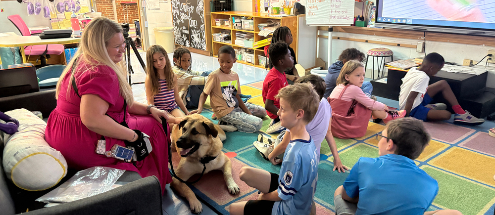 Women with blonde hair and pink dress hold leash with light brown dog next to 11 kids sitting on floor.