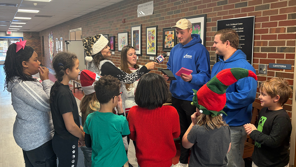 A group of elementary students wearing festive holiday hats gather in the hallway as a FOX 4 reporter stands next to School Clerk Kristina Peister and holds a microphone toward two men in blue hoodies from Accountable Mudjacking.