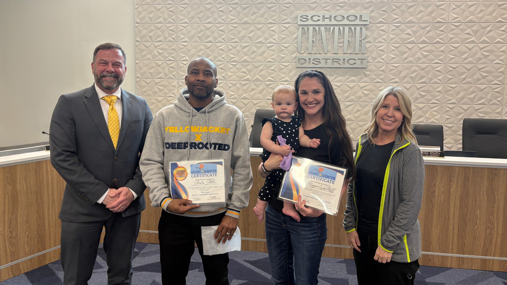 Superintendent Dr. Troy Hogg, Heart of Center recipients Andre Allen and Morgan Westhoff, and Board of Education Chair Marcie Calvin stand in front of the board dias.