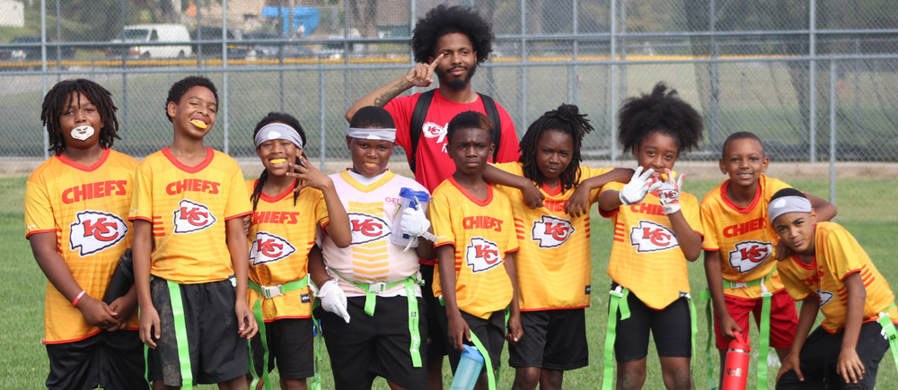Nine kids in yellow t-shirts with KC Chiefs on the front stand on a green football field.