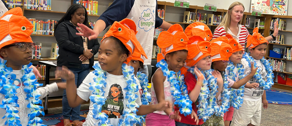 Kids in clownfish hats and flower leis  perform in front of a bookshelf.