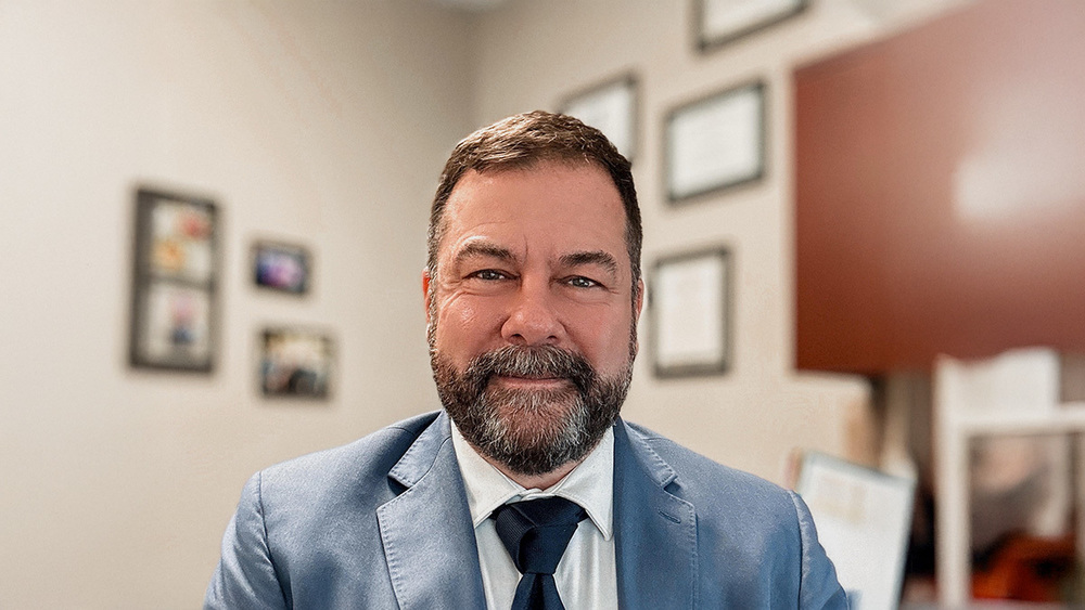 Man in a light-gray suit and dark tie, smiling and looking directly at the camera.