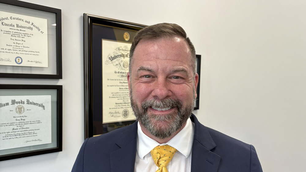 Dr. Troy Hogg stands in front of his diplomas in a blue suit and yellow tie.