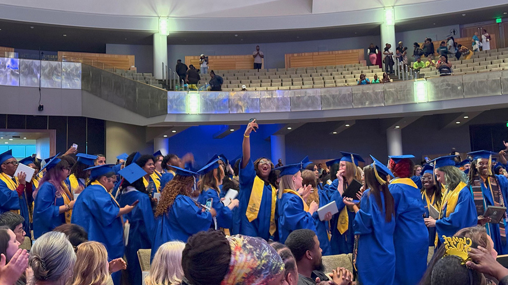 Graduates in blue gowns and yellow stoles celebrate joyfully in a large auditorium. They hold diplomas and smartphones, capturing the moment.