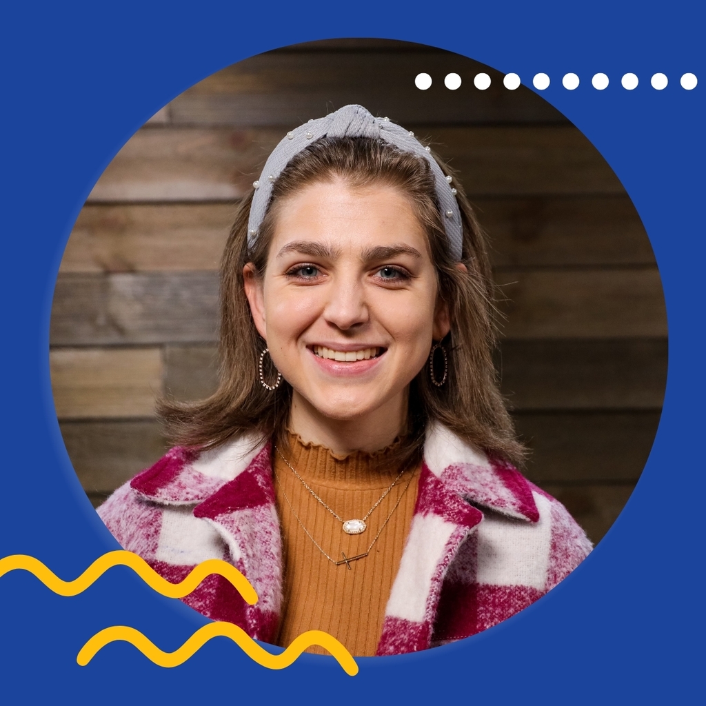 Woman with brown hair, a headband, and red checkered jacket stands in front of a wood wall, smiling.