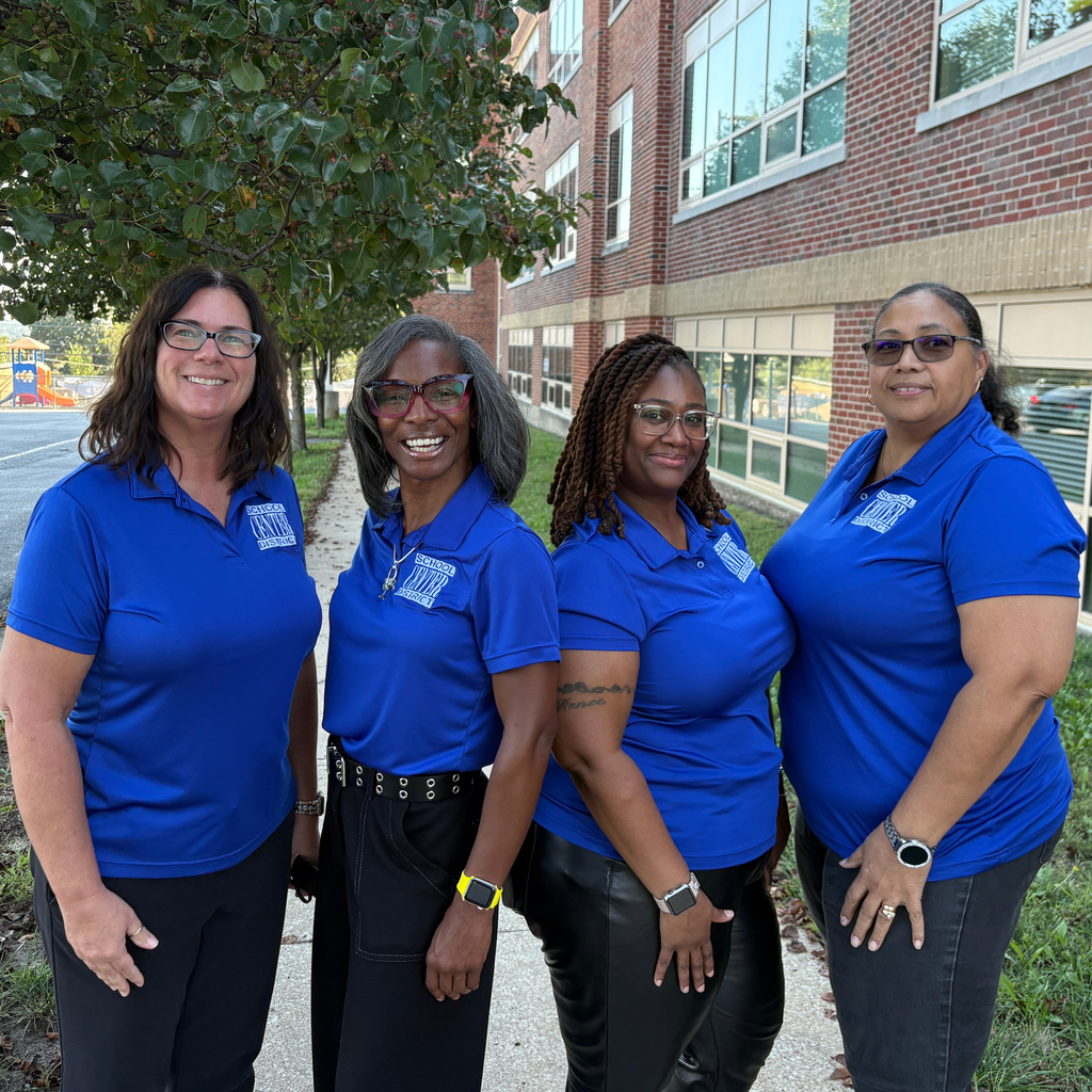Four women in matching blue collared shirts and black pants stand in front of a brick building.