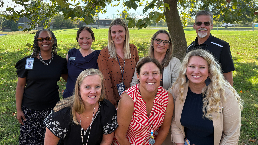 Four women and one man stand in a row behind 3 women. hands on their knees with a green grassy field in the background.