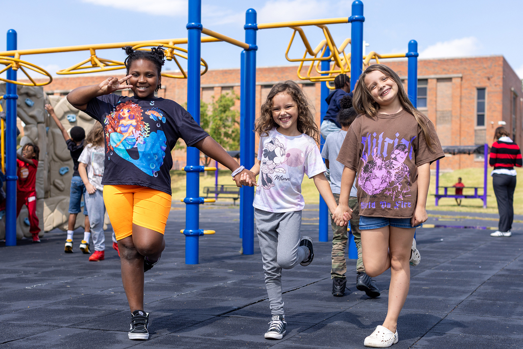 Three girls stand on the playground holding hands and smiling.