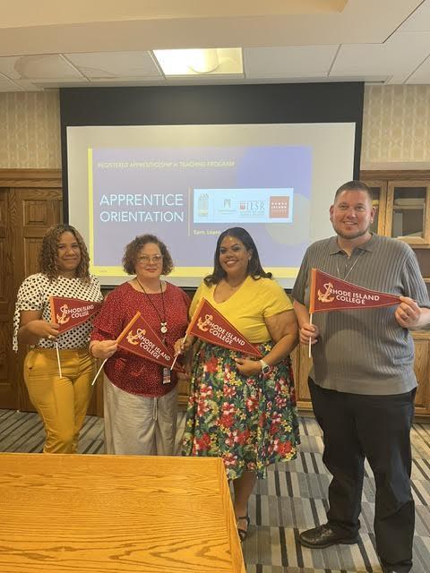 Four individuals standing together indoors during an apprenticeship orientation event, each holding a red Rhode Island College pennant. A presentation screen in the background displays the words “Apprentice Orientation” along with organization logos. The group is smiling and posing in a conference or training room setting.