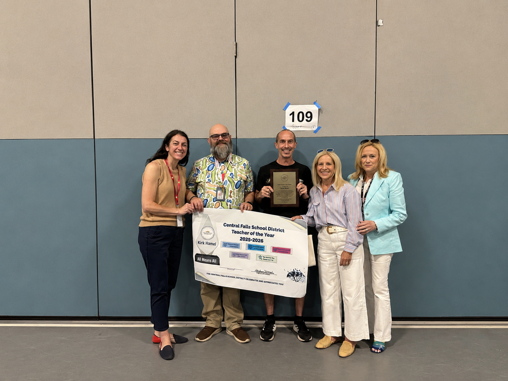 From left to right, Superintendent Dr. Stephanie Downey Toledo, Veterans Memorial Elementary School Principal Mike Carline, 2025–2026 Central Falls School District Teacher of the Year Kirk Hamel holding his award plaque, Chief Academic Officer Joy Souza, and Deputy Superintendent Ann Lynch pose together in a school gymnasium. The group holds a large Central Falls School District Teacher of the Year 2025–2026 recognition banner honoring Kirk Hamel. A gym wall and room number sign are visible in the background.