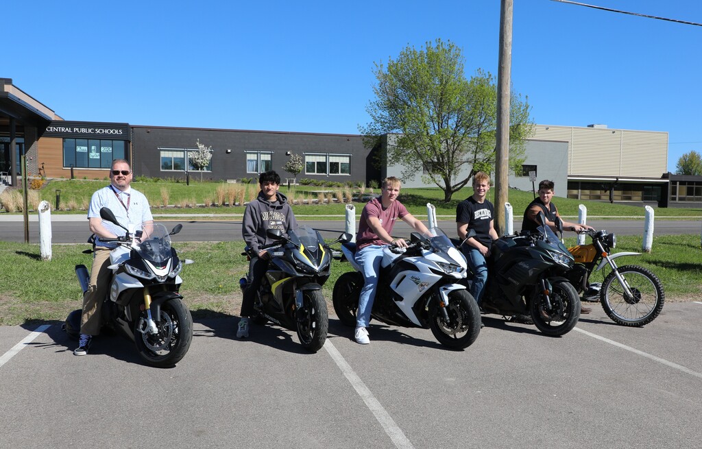 4 students and one teacher sitting on motorcycles they drove to school.
