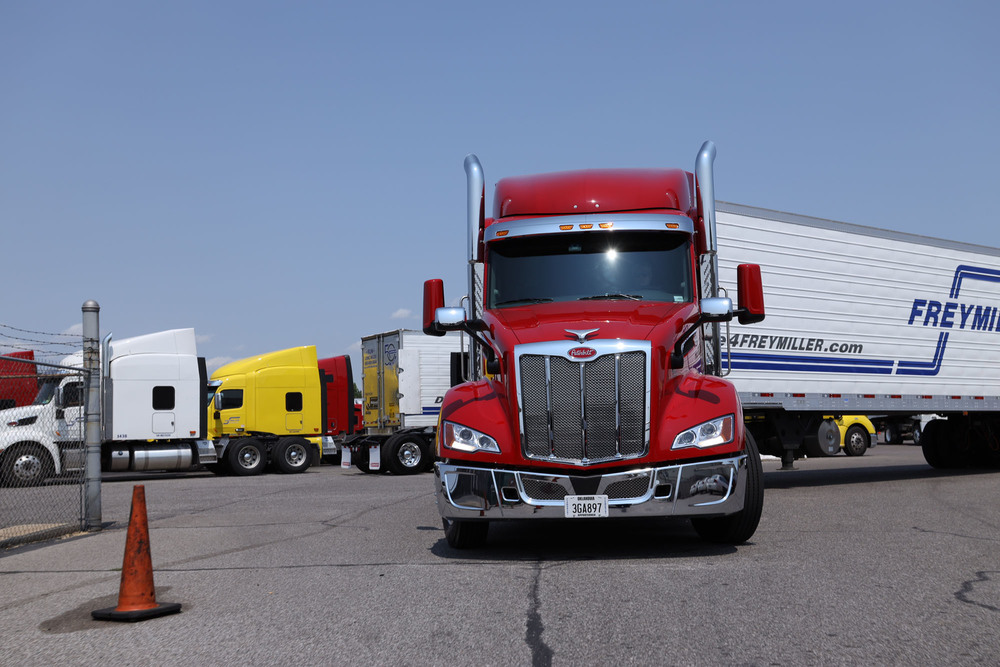 A bright red Peterbilt semi-truck is parked in a paved lot under a clear blue sky. It is hitched to a white trailer with the Freymiller company logo. Other white and yellow trucks are visible in the background.