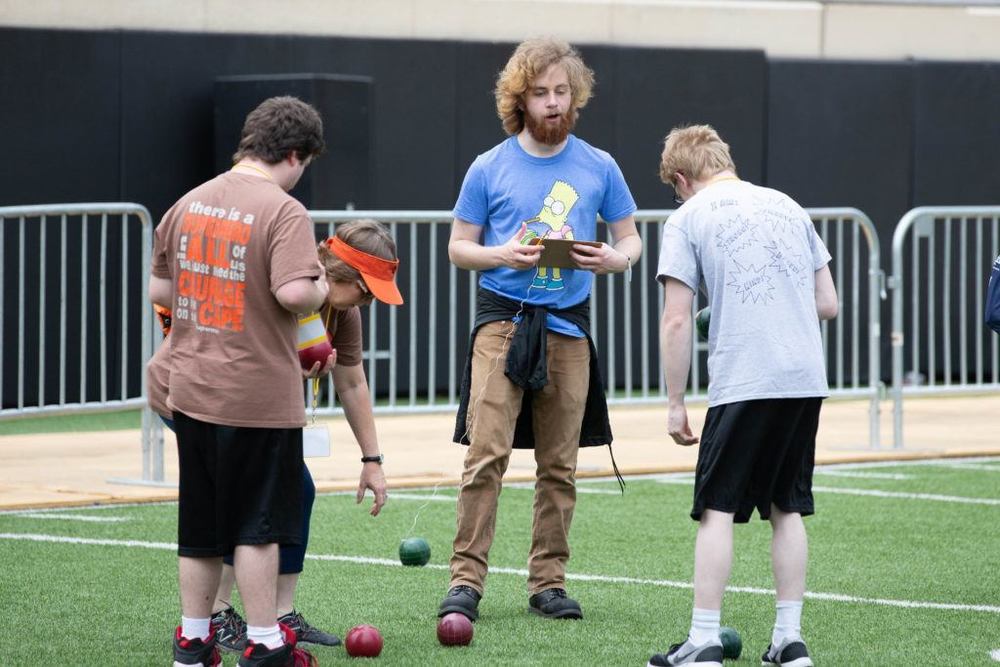 Four people play bocce on a turf field. A bearded man in a blue shirt holds a clipboard and string to measure distance between the balls.
