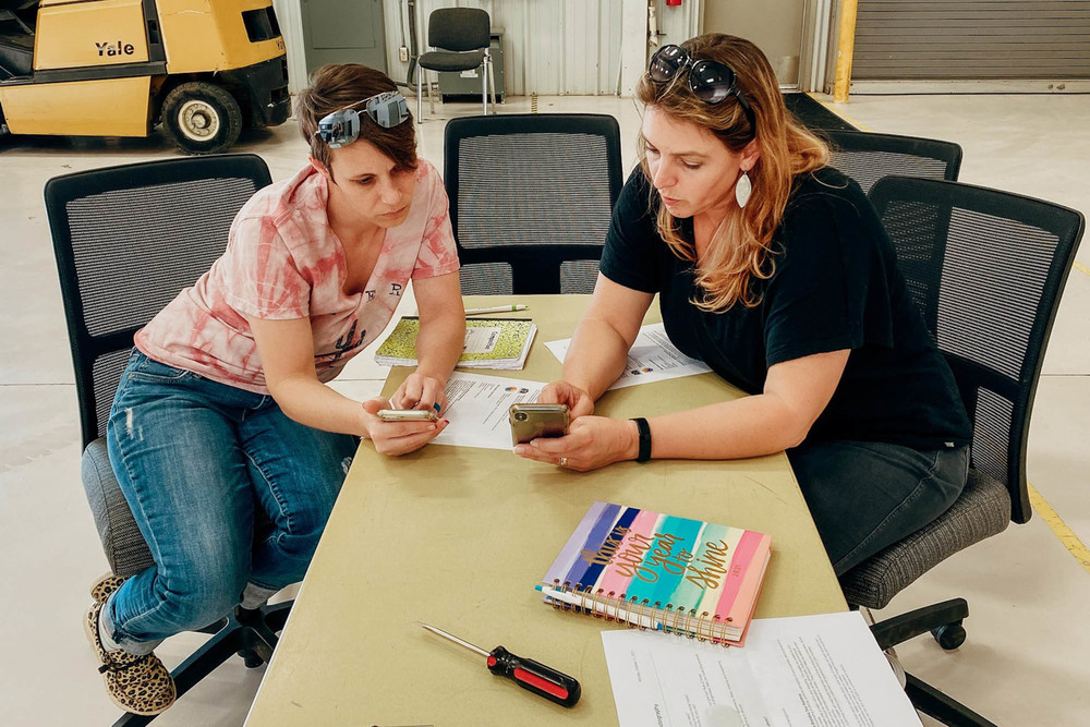 Two women collaborating at a table in a bright industrial workspace, using their phones to review information.