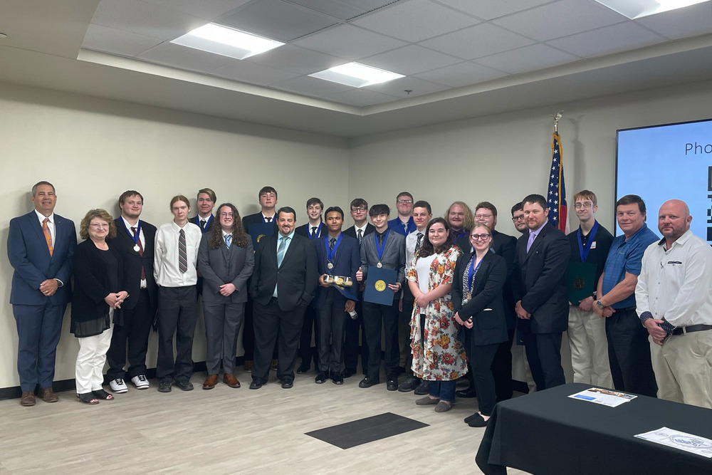 A large group of young adults and several older professionals pose for a formal group photo in a conference room. Many of the young men wear suits and ties, with some sporting blue ribbons and medals around their necks. An American flag stands in the background next to a digital display.