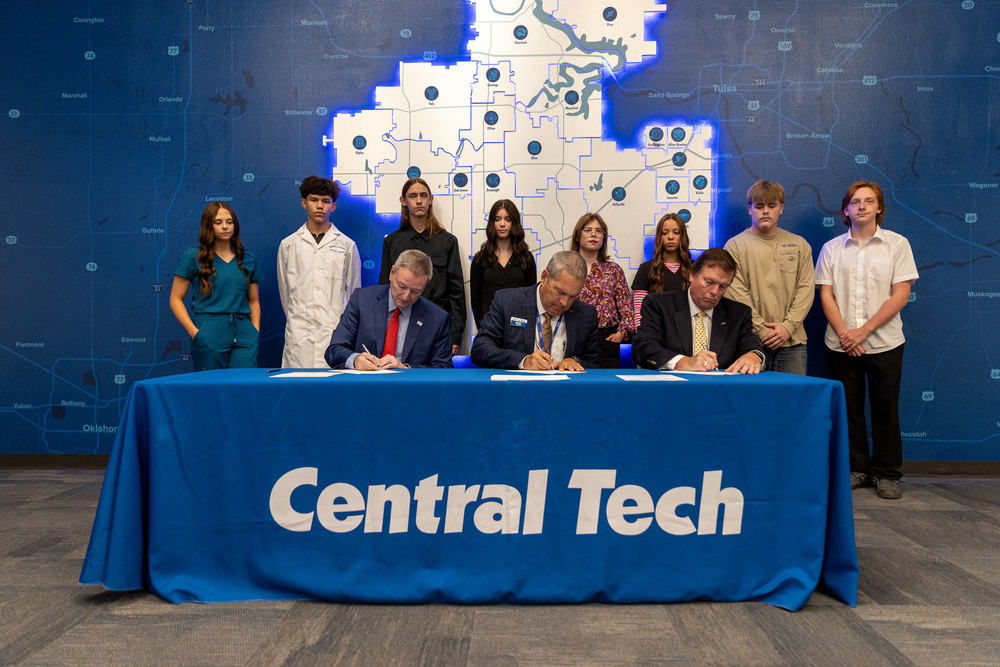 The Rogers State University President sits at a table with the Central Tech Superintendent and Assistant Superintendent as the sign the agreement and 8 students stand behind them.