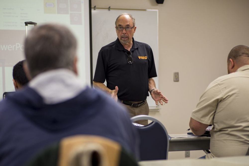 A male instructor in a black polo shirt and glasses gestures with open hands while teaching a seated class with a projector behind him.