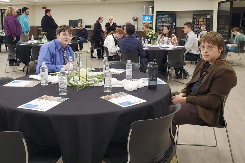 Two students sit at a round table with a black tablecloth during a formal event. The table is set with bottled water and informational flyers. Other guests are visible in the background near a snack area.
