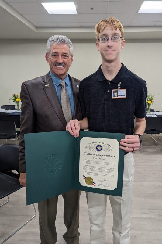 A young man with glasses and a black polo shirt holds a green "State of Oklahoma Citation of Congratulations" for Ryan Chestnut. He stands alongside a smiling man in a brown leather blazer.