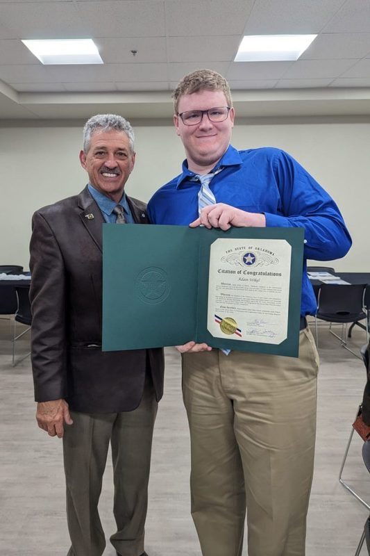 A young man in a blue button-down shirt and glasses smiles while holding open a green "State of Oklahoma Citation of Congratulations" folder. He is standing next to an older man in a brown leather blazer and tie in a banquet hall.