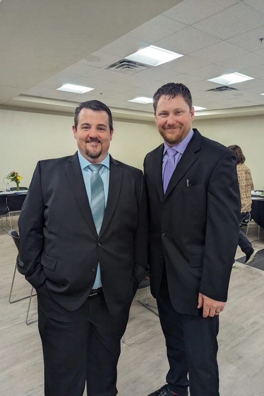 Two men in black suits and ties stand side-by-side smiling for a photo in a banquet hall. The man on the left wears a light blue tie, and the man on the right wears a purple tie.