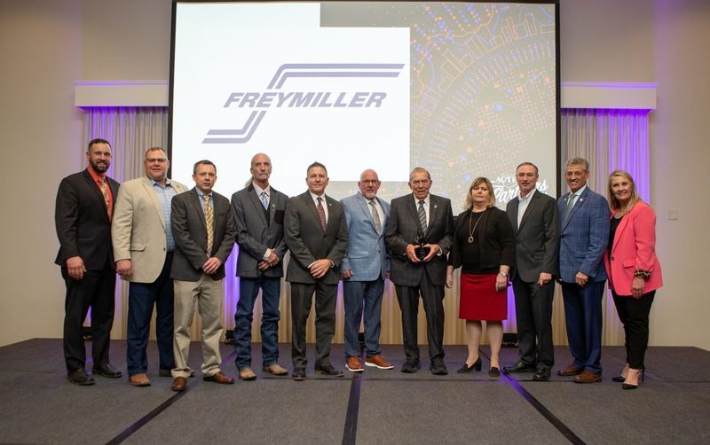 A group of eleven professionals in business attire stand in a line on a stage for a formal photo. Behind them, a large projector screen displays the Freymiller company logo. The man in the center holds a small black award trophy.