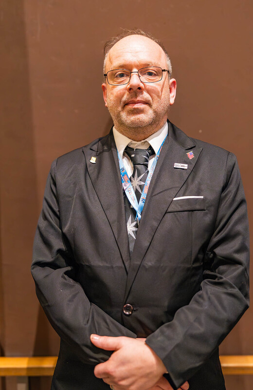 man wearing glasses in a suit jacket standing in front of a beige walls with his hands crossed