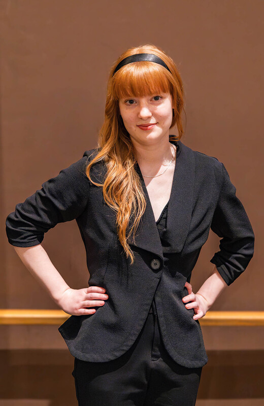 young woman with red hear wearing a black headband and blazer with her hands on her hips in front of a beige wall