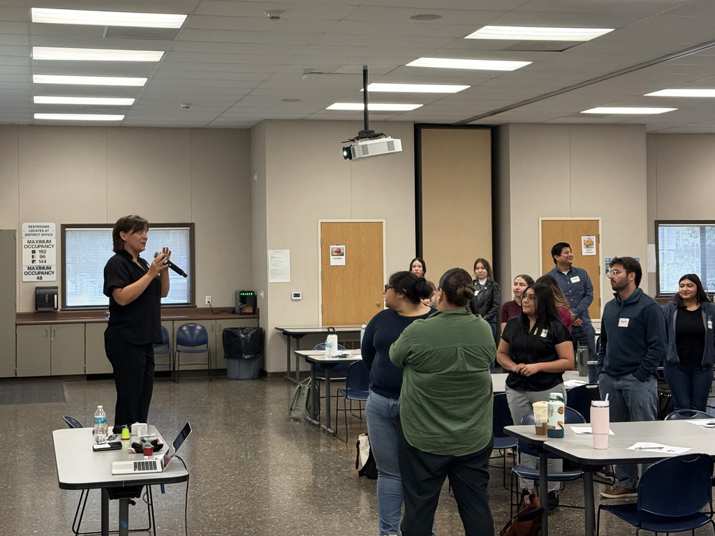 Presenter, holding microphone, addresses standing teachers