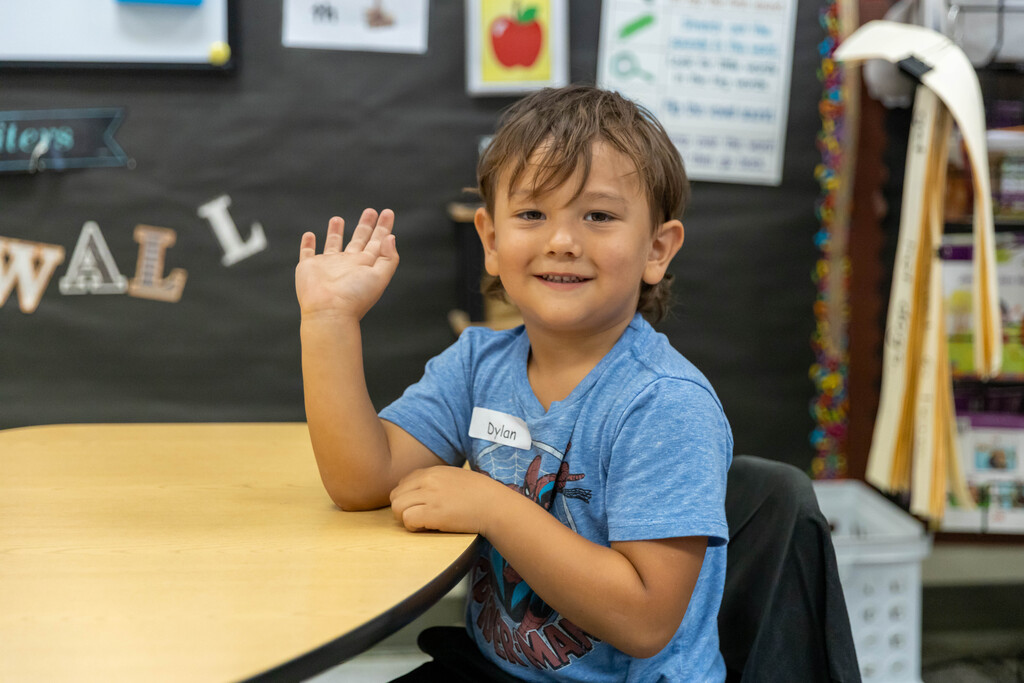 Young student sitting at table waves and smiles