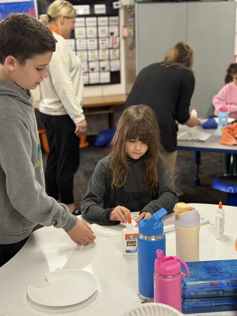 Sixth grade students sit beside first graders, helping them create Rainbow Fish art projects after reading the book together in Mr. Wise’s classroom.