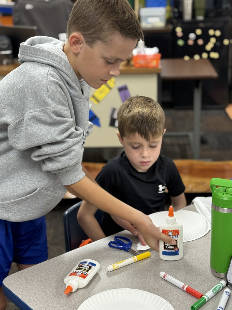 Sixth grade students sit beside first graders, helping them create Rainbow Fish art projects after reading the book together in Mr. Wise’s classroom.