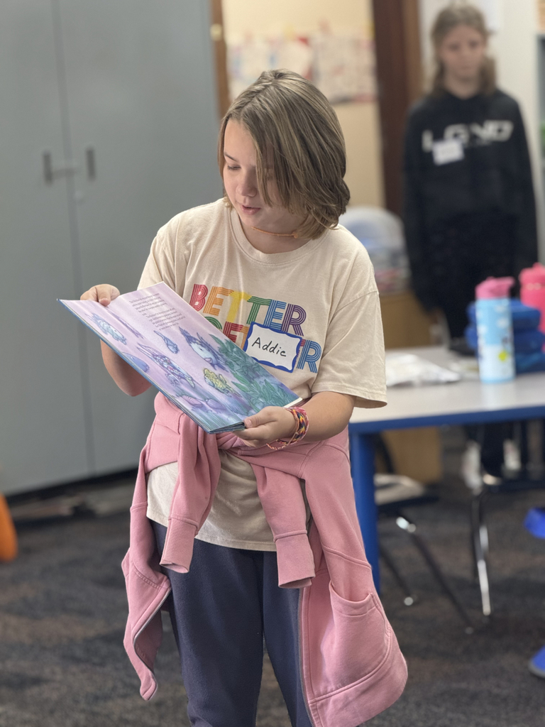 Sixth grade students sit beside first graders, helping them create Rainbow Fish art projects after reading the book together in Mr. Wise’s classroom.