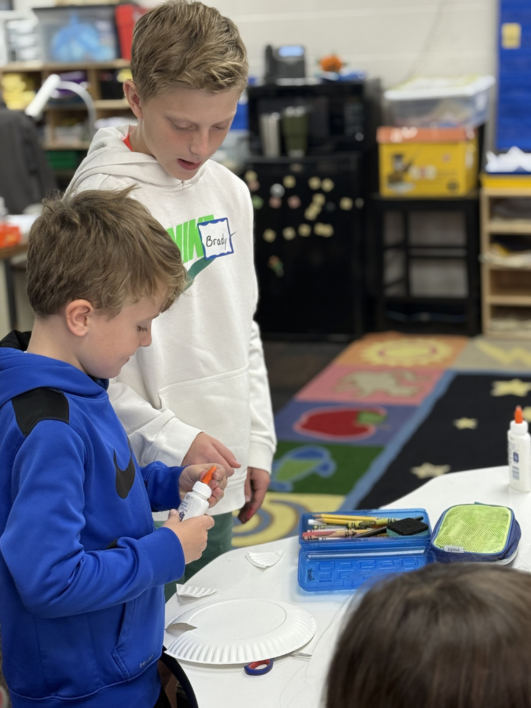 Sixth grade students sit beside first graders, helping them create Rainbow Fish art projects after reading the book together in Mr. Wise’s classroom.